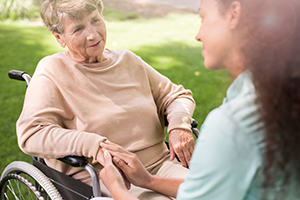 young nurse kneeling down in front of elderly woman in wheelchair holding her hand